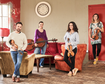 The Elias String Quartet holds their instruments in a living area with red accents decorating the room.