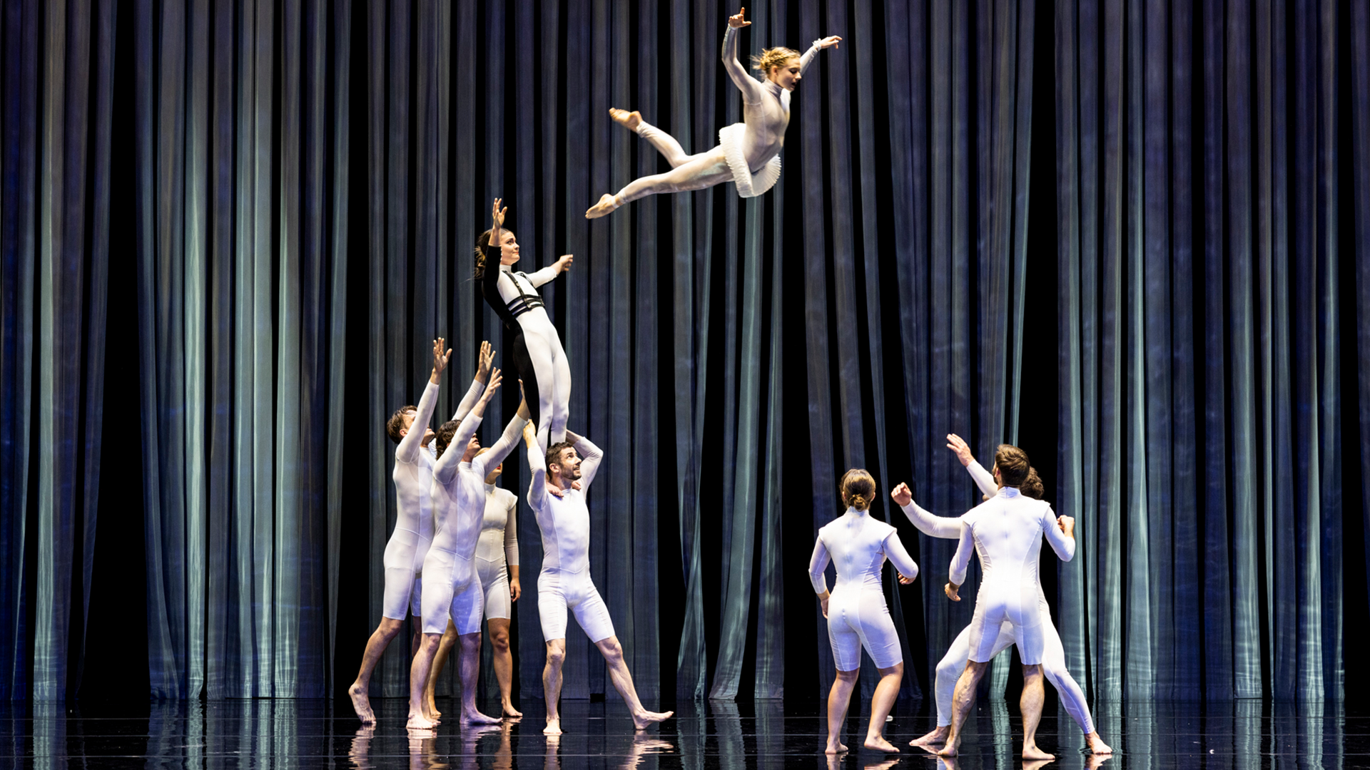 Dynamic photo of acrobats from Circa Contemporary Circus mid-performance, dressed in white unitards. One performer is soaring through the air above a group of others with arms outstretched, set against a dramatic, dark vertical-striped stage backdrop.