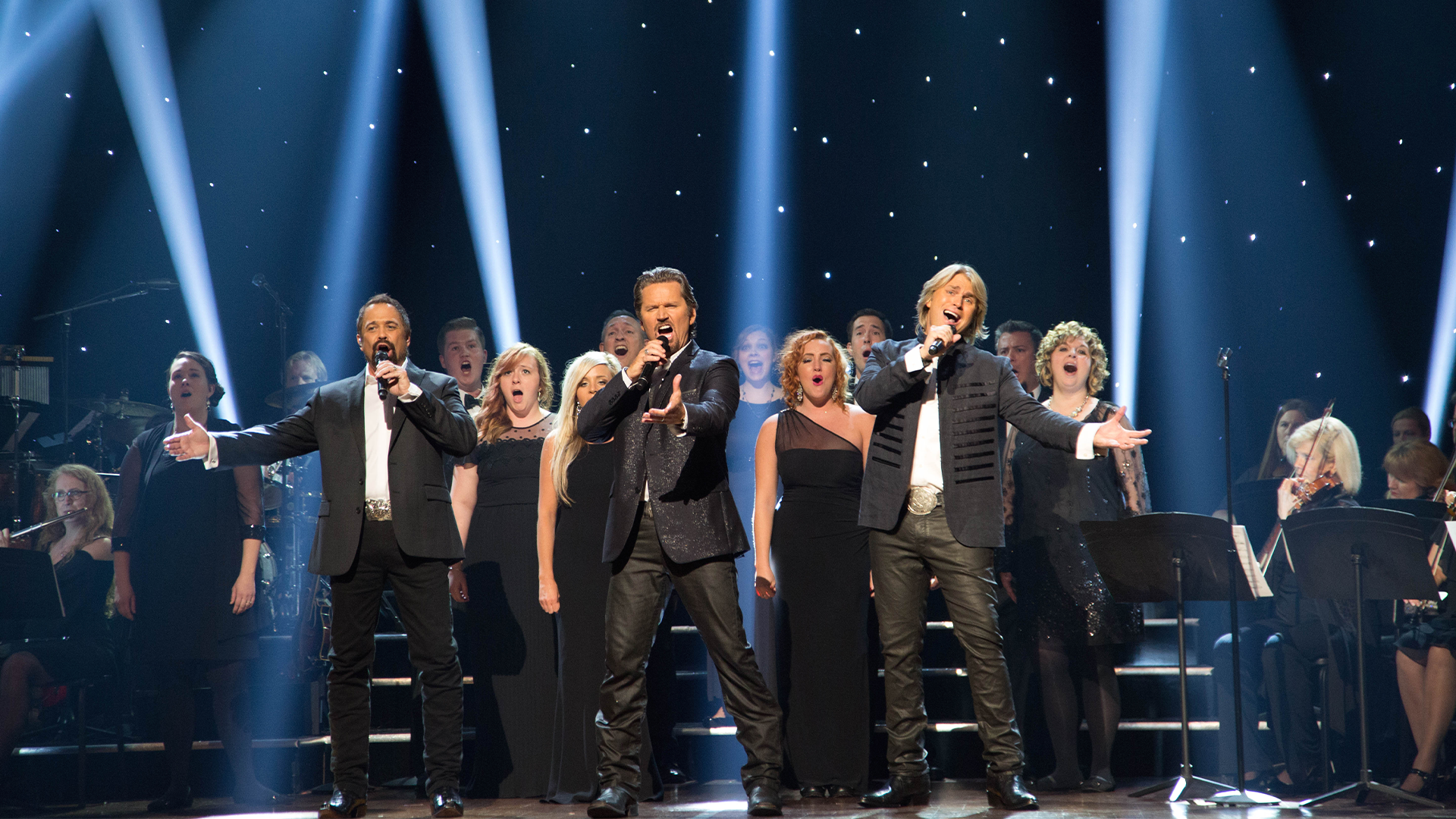 A group of men singing in black suits on stage with backup singers