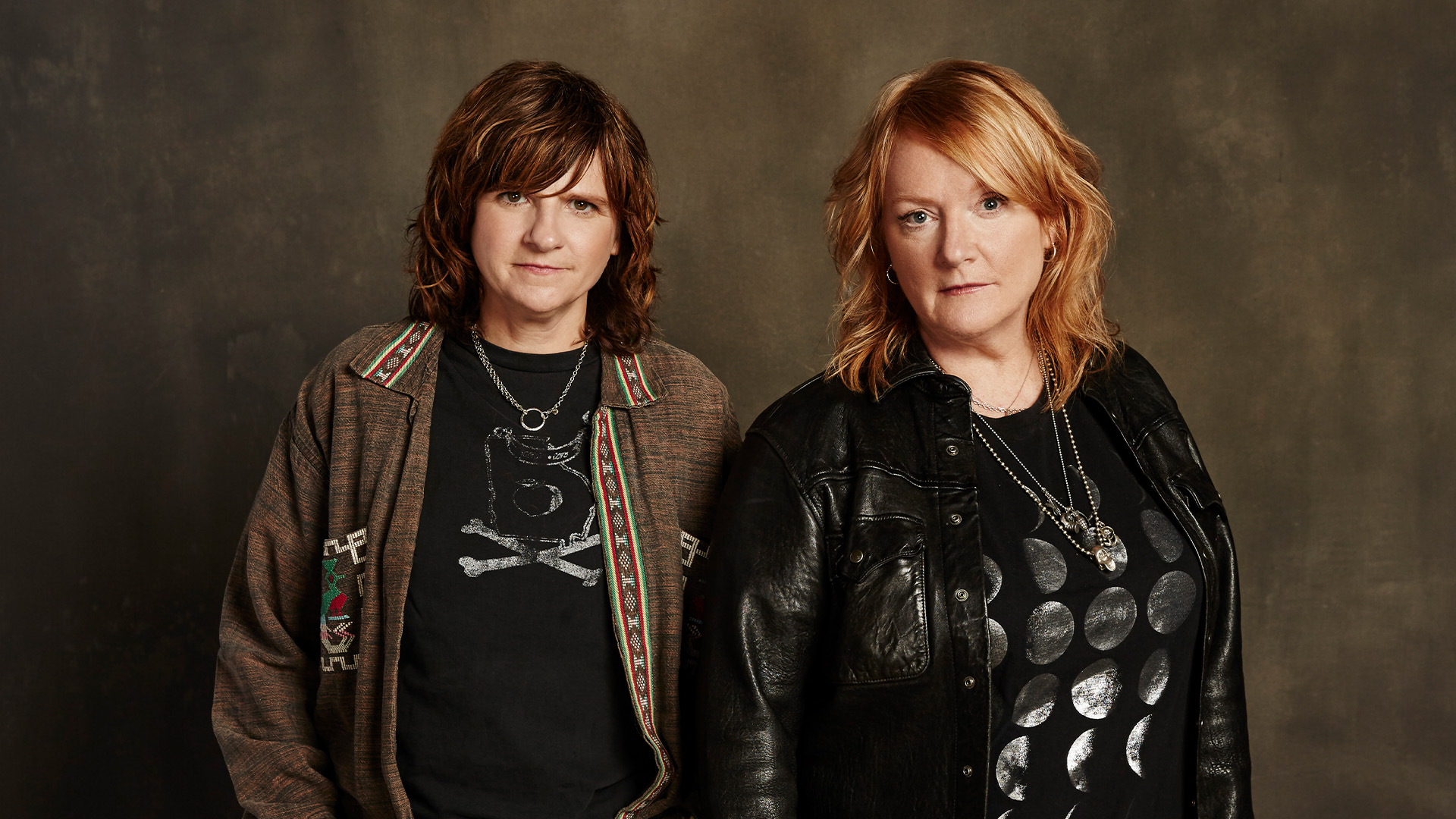 Two people dressed in black look at the camera with necklaces on. 