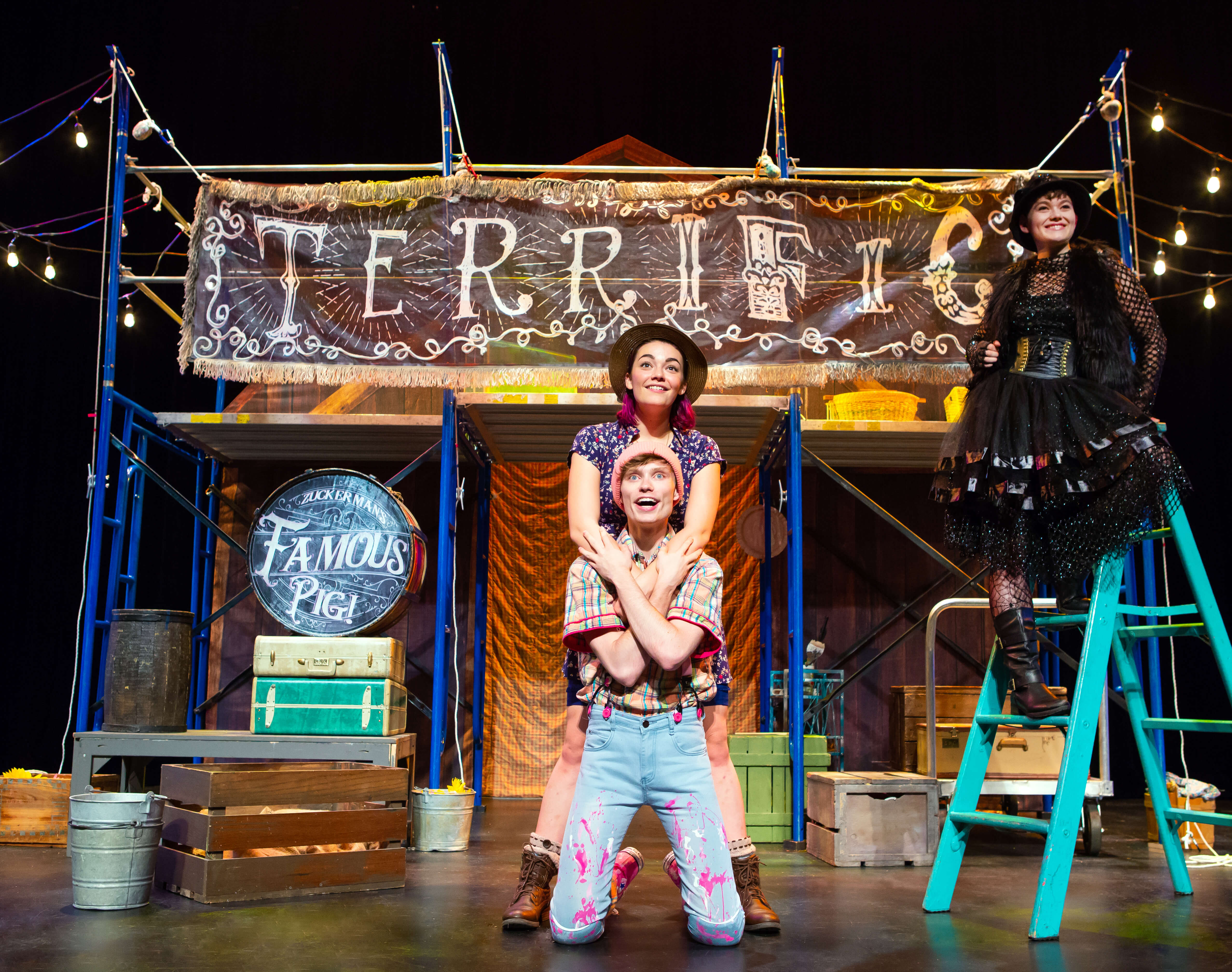 Two people on stage holding each other while another actor in all black stands on a ladder and smiles. Setting is a fairground