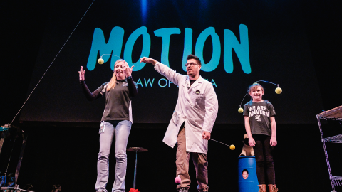 A man in a lab coat and two people on stage with tennis ball floating in the air 