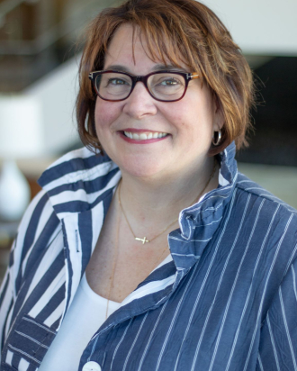 A headshot of Camie Gunderson wearing a blue striped shirt and brown glasses.