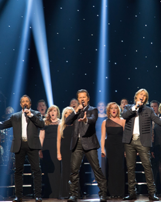 A group of men singing in black suits on stage with backup singers