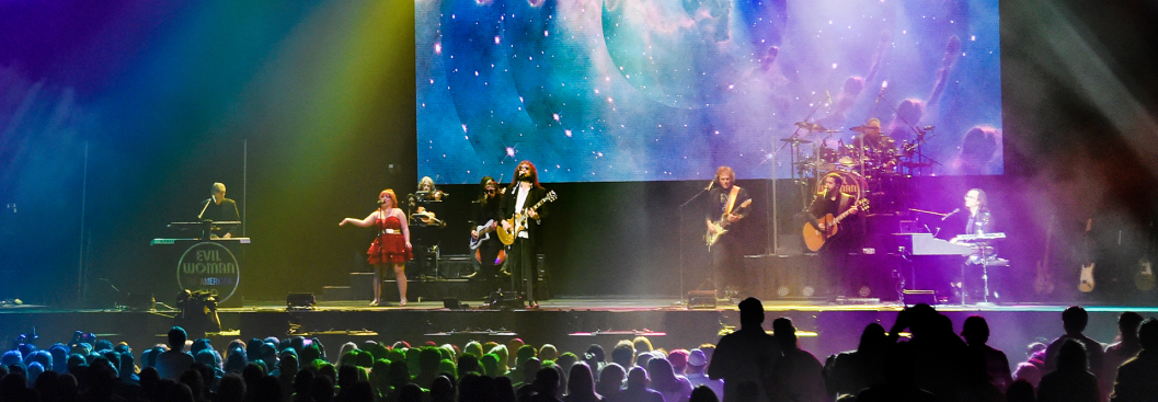 A hazy stage illuminated by moody, multi-colored lights with 12 musicians performing on a blend of rock and classical instruments