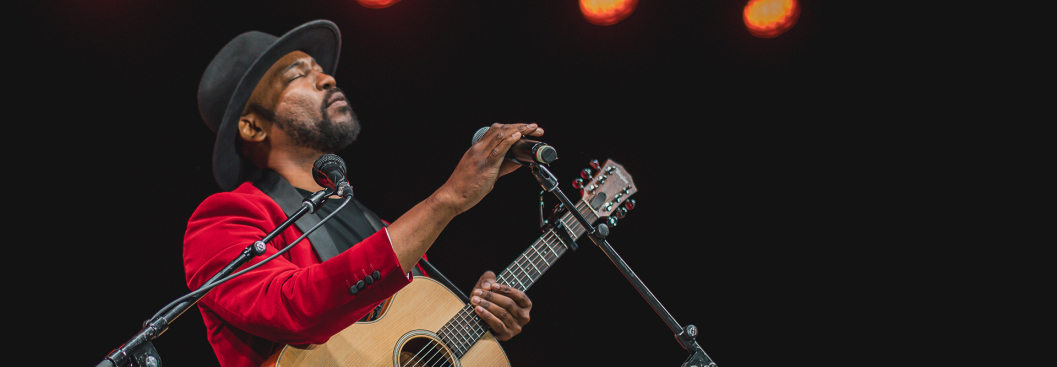 Musician Rocky Peter stands on stage, eyes closed while singing into a microphone and holding an acoustic guitar