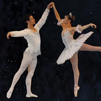 A pair of male and female ballet dancers dressed in all white pose together with pointed toes